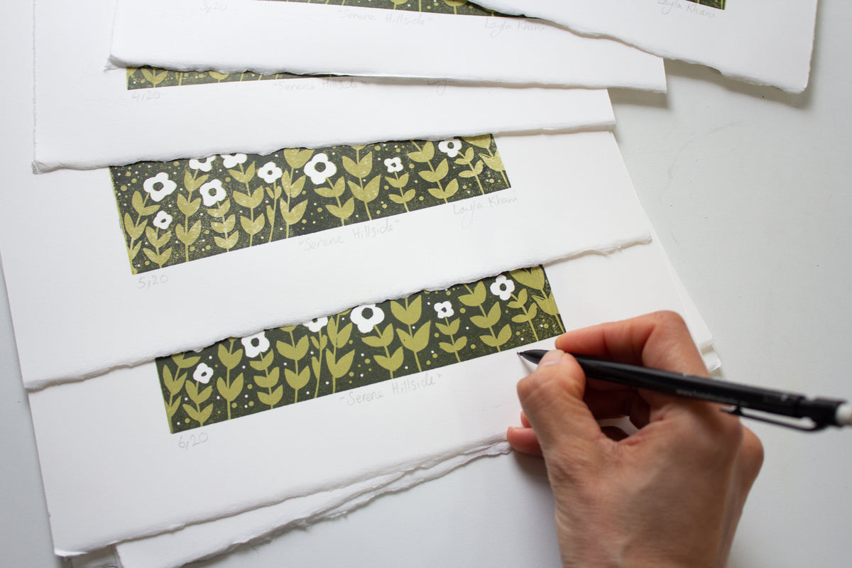 Artist signing and numbering a small limited-edition reduction linocut print titled Serene Hillside, surrounded by hand-printed editions on deckled white paper.