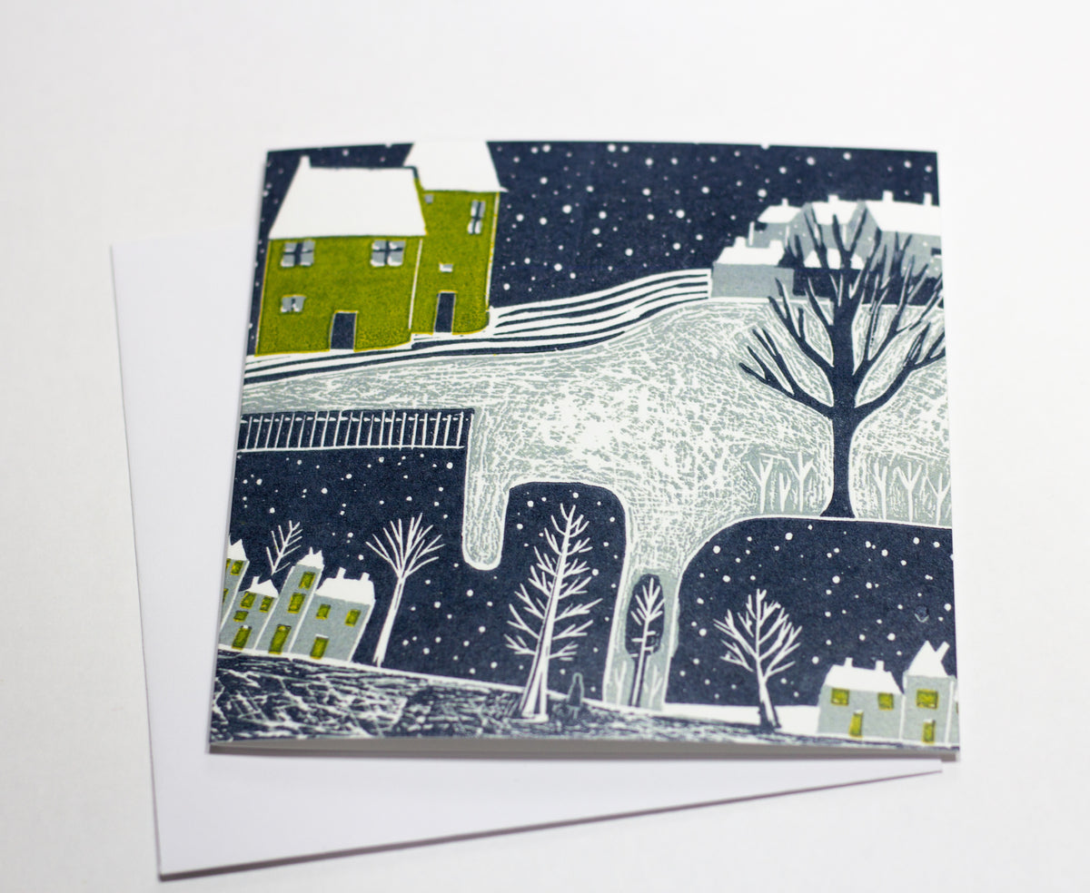 Wintry Night Christmas linocut greeting card featuring snow-covered cottages, a green house, bare trees and a starry winter sky, photographed with its white envelope.