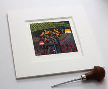 Load image into Gallery viewer, A colourful linocut print titled "Autumn Glow" shows an autumn tree, hills, a white house, and red plants. The artwork is framed and placed on a white surface, with a lino cutting tool nearby. The edition number and artist's signature are underneath.