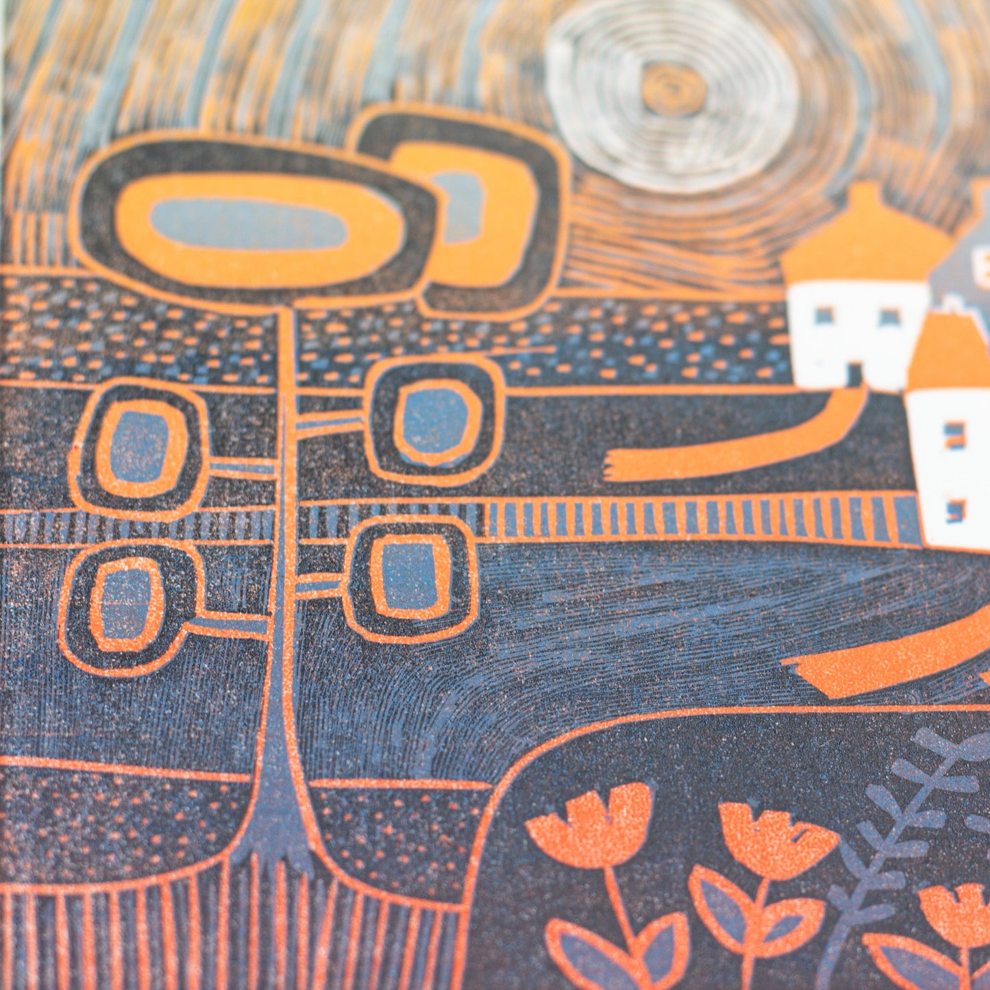 Close angled view of an original mini linocut print, white houses and trees under a swirling orange moon