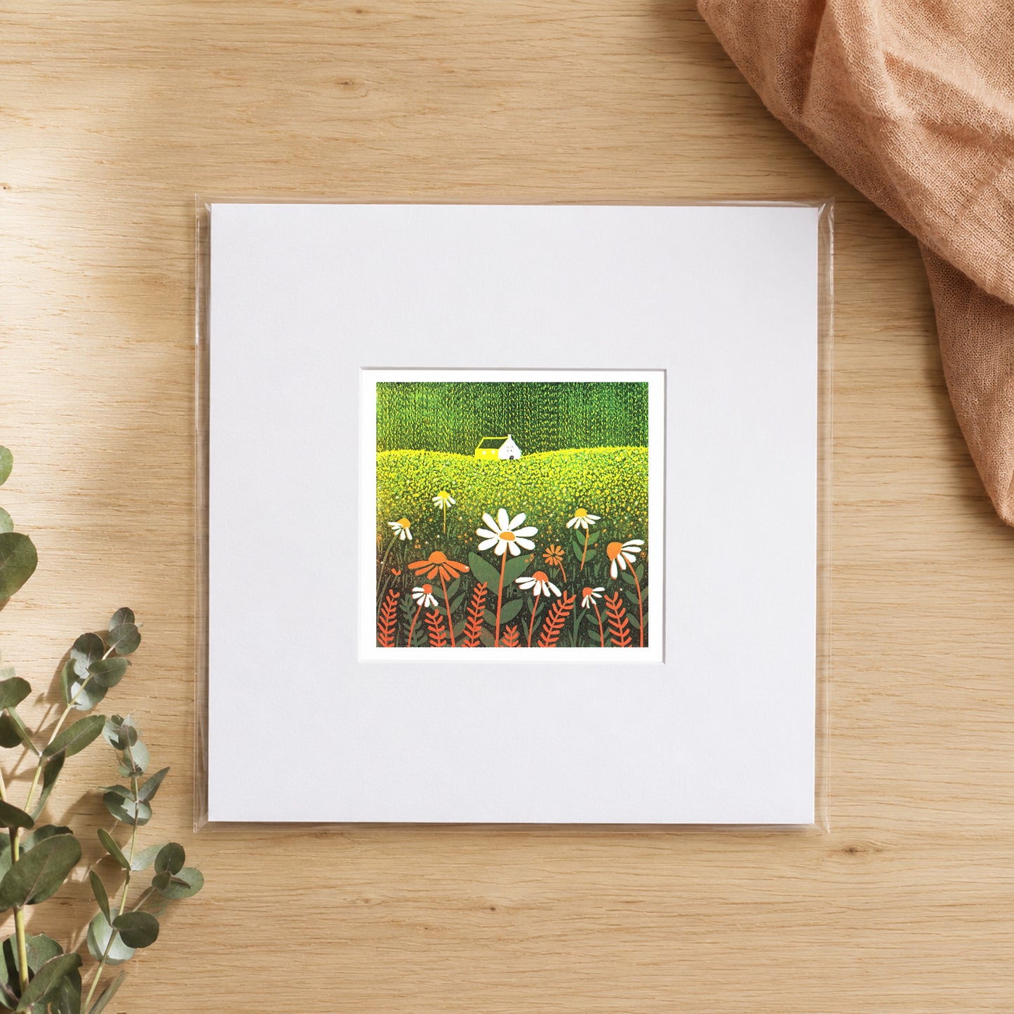 Mini linocut daisy meadow print in a white window mount, photographed flat on warm oak with eucalyptus