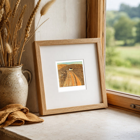 Mini linocut print framed in pale oak, propped on a windowsill with dried wheat in a stone vase