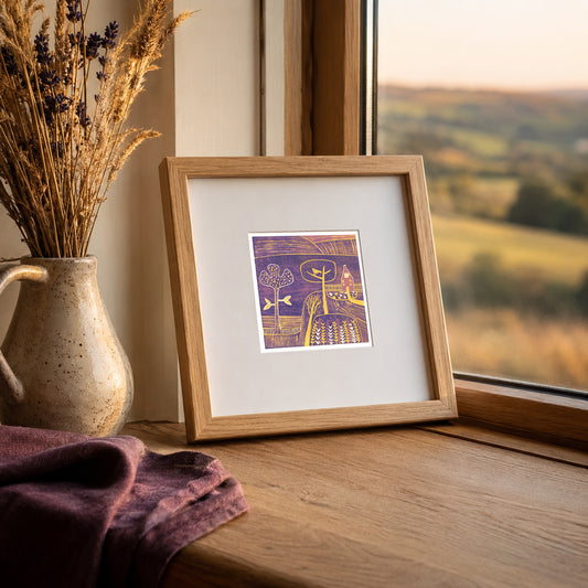 Mini linocut print framed in pale oak, propped on a windowsill with dried grasses in a stone vase