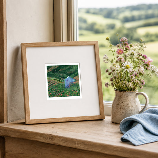 Mini linocut print framed in pale oak, propped on a windowsill with dried stems in a stone jug
