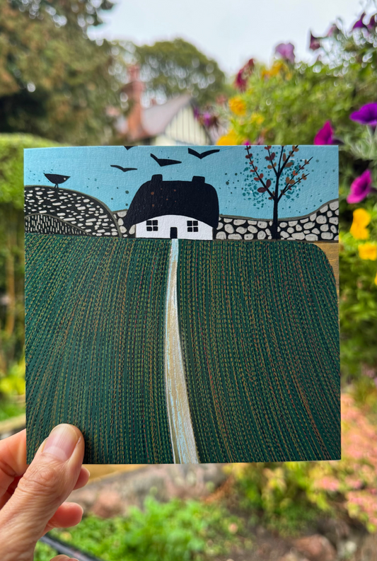 Hand holding a square linocut greeting card featuring a white cottage, stone walls and green fields, photographed outdoors against a colourful garden.