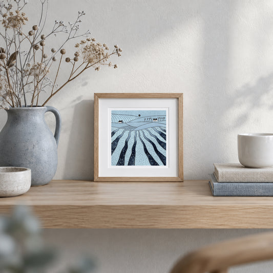 Where the Hills Breathe blue landscape linocut displayed in a natural wood frame on a shelf beside a ceramic vase of dried flowers.