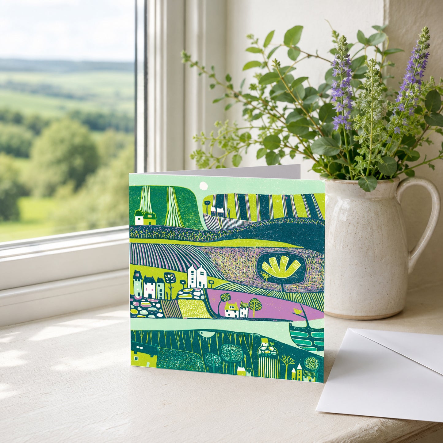 Linocut folk village greeting card propped on a windowsill with greenery, overlooking the hills