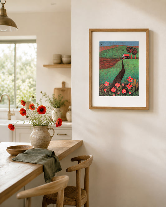 Framed The Red Roofed Cottage linocut print displayed in a warm dining area with natural wood furniture and flowers.