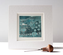 Load image into Gallery viewer, linocut print titled 'Serene Paths' by Layla Khan, showcasing a stylized countryside landscape with trees, rolling hills, and a small house in soft blue tones. The print is numbered 3/20 and rests against a light background, accompanied by a wood-handled lino carving tool in the foreground.