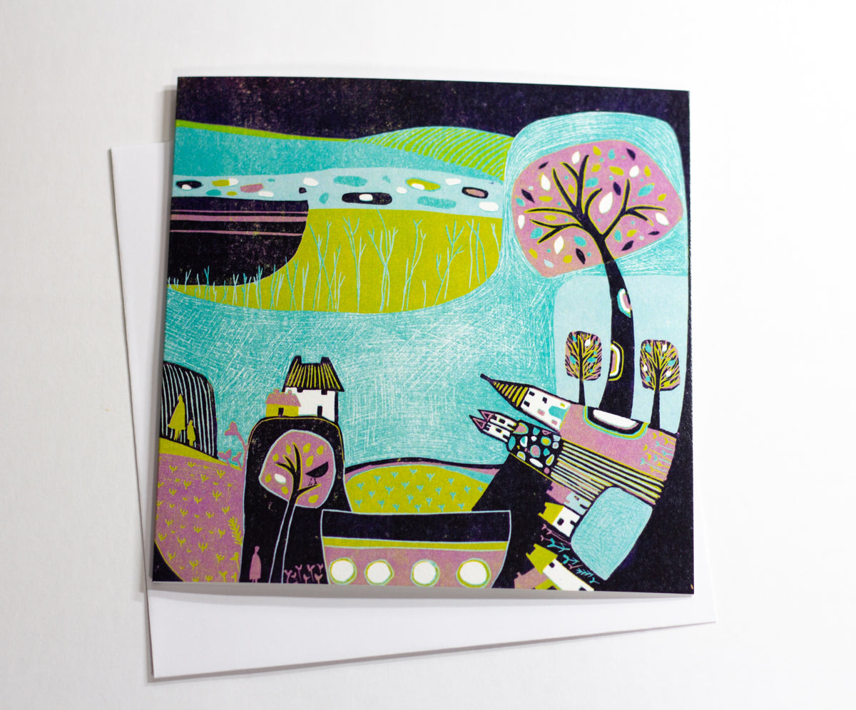 Square linocut greeting card featuring a colourful countryside scene with turquoise fields, a pink tree and cottages, photographed against a white background.