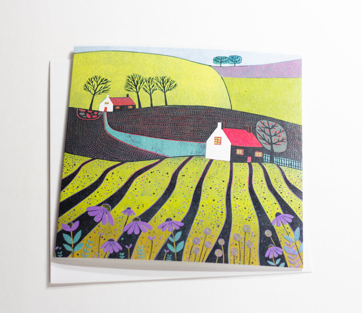 A Glorious Spring Day linocut greeting card featuring bright green countryside, white cottages, rolling fields and purple wildflowers, photographed with its white envelope.