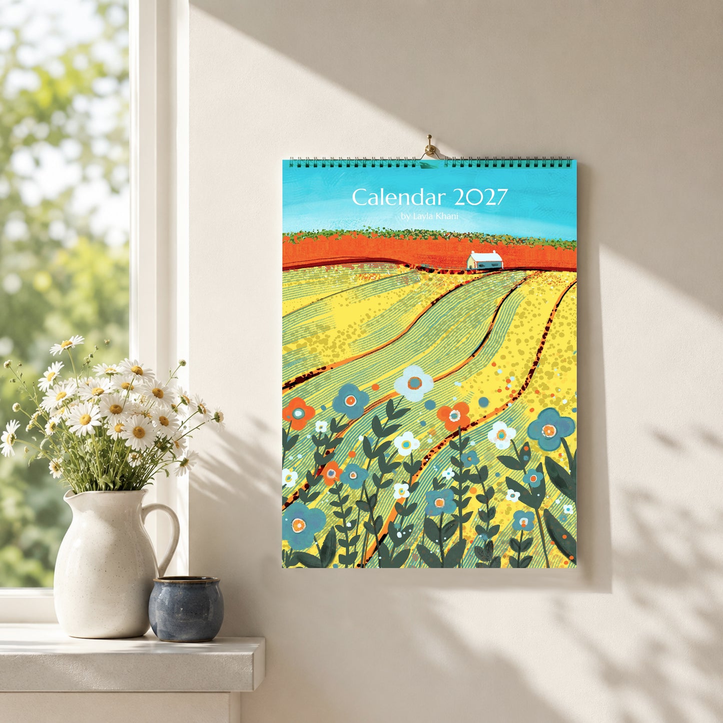 A4/A5 2027 art wall calendar with colourful countryside cover, hanging in a bright neutral interior beside flowers and ceramics.