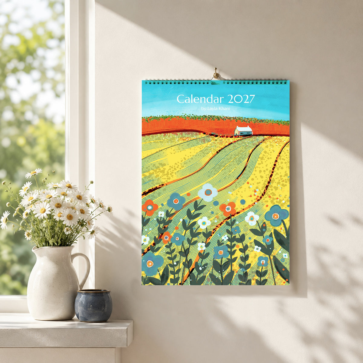 A4/A5 2027 art wall calendar with colourful countryside cover, hanging in a bright neutral interior beside flowers and ceramics.