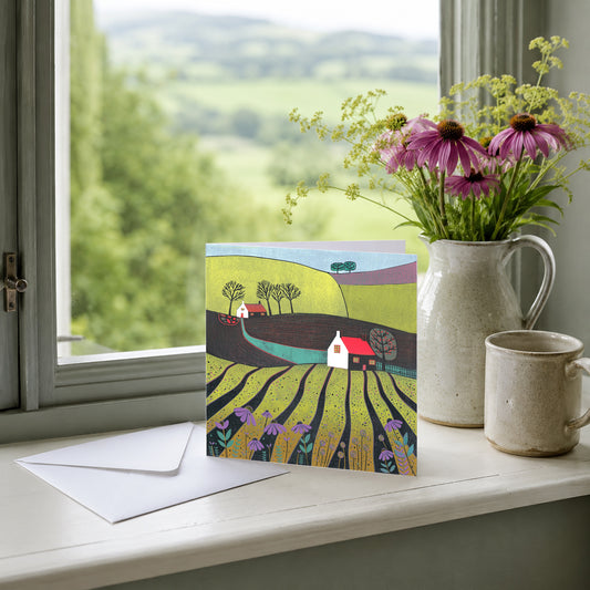 A Glorious Spring Day linocut greeting card featuring colourful green fields, a cottage and trees, photographed in a lifestyle setting.