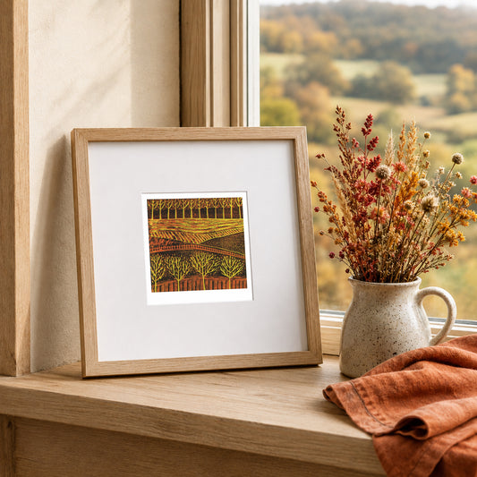 Mini linocut print framed in pale oak, propped on a windowsill with dried stems in a stone jug