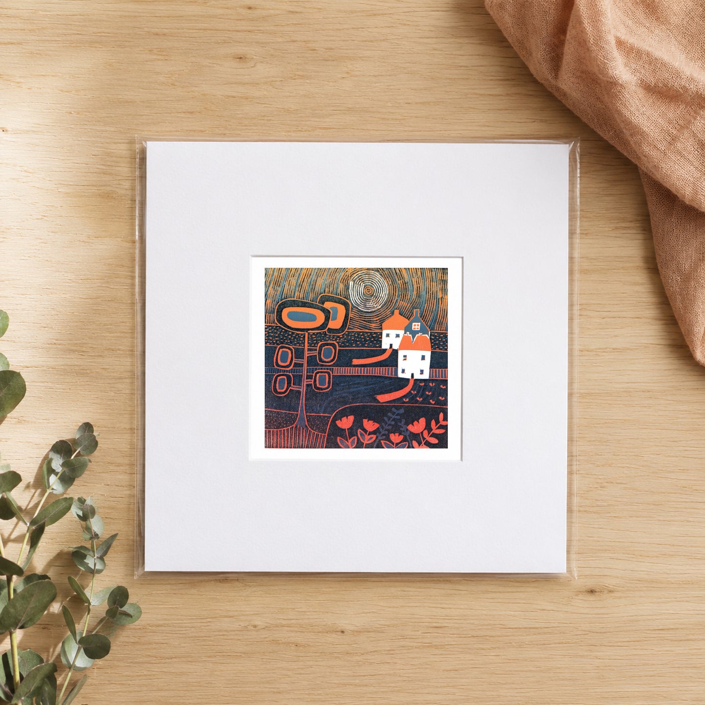 Mini linocut moonlit village print in a white window mount, photographed flat on warm oak with eucalyptus