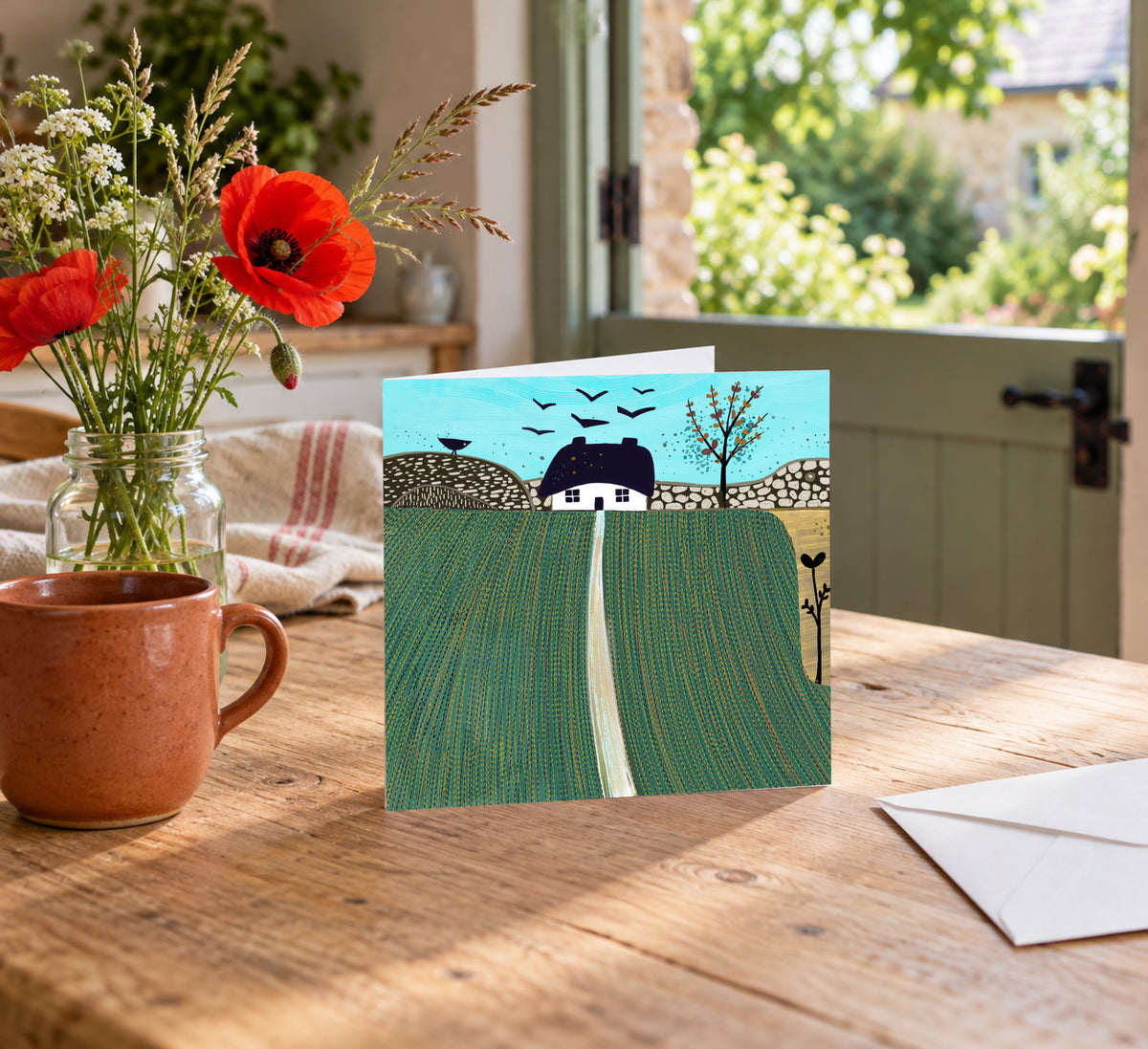 Autumn on the Hills square greeting card displayed on a wooden table beside a ceramic vase of flowers, with a garden visible through the window.