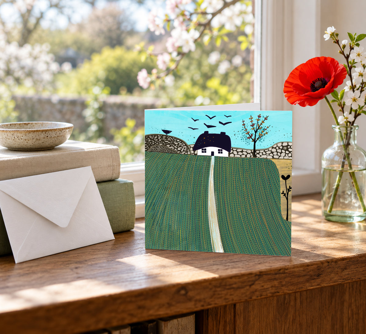Autumn on the Hills greeting card displayed on a wooden table beside a vase of red flowers, featuring green fields, a white cottage and a turquoise sky.