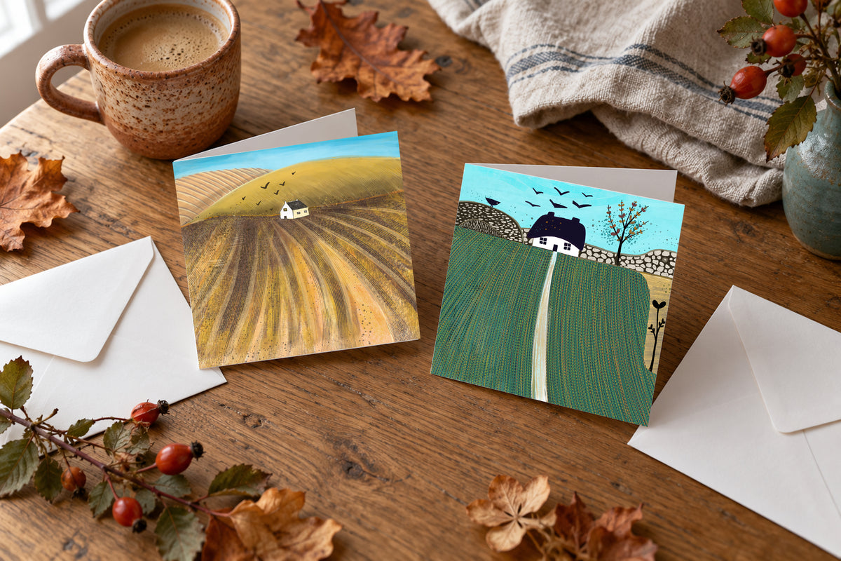 Four countryside art greeting cards in two autumn landscape designs, arranged on a wooden table with envelopes, a ceramic mug and autumn leaves. Copy