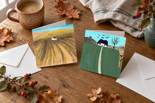 Four autumn countryside greeting cards in two landscape designs, arranged on a wooden table with envelopes, a ceramic mug and autumn leaves.