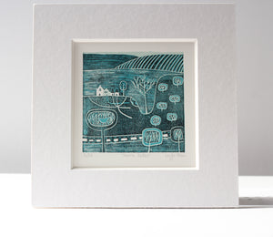 A detailed linocut artwork titled 'Serene Paths' by Layla Khan, featuring a tranquil rural scene with minimalist trees, winding paths, and a farmhouse. The print, in cool blue hues, is framed in a white mount, with a lino cutting tool placed beside it for a touch of the creative process.