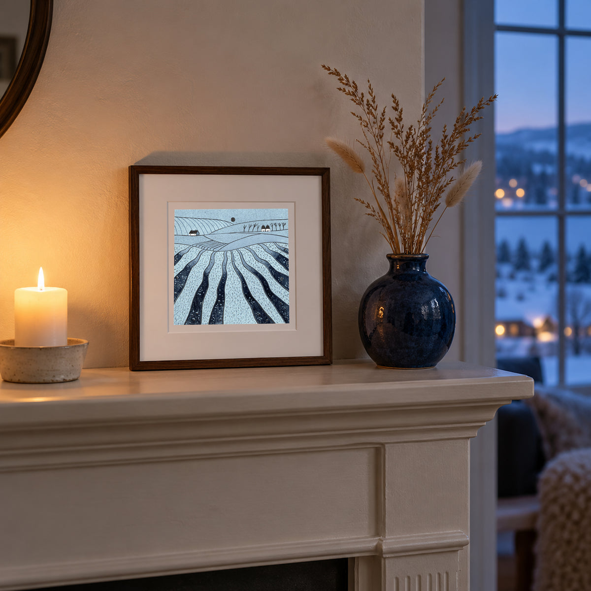 Where the Hills Breathe linocut print in a wooden frame on a mantelpiece beside a lit candle and vase, with a winter evening view.
