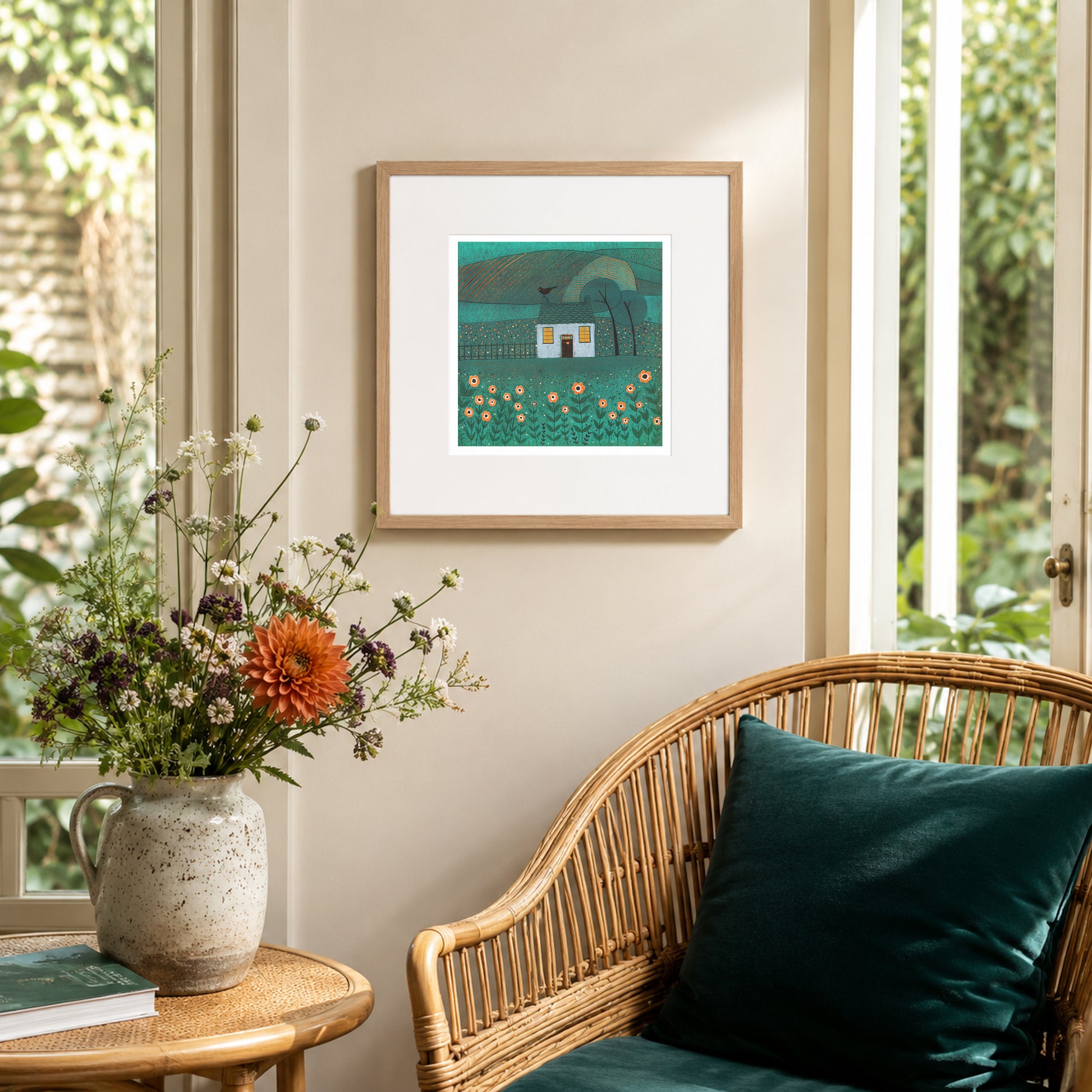Framed The House with the Bird linocut print displayed in a bright garden room with a rattan chair and flowers.