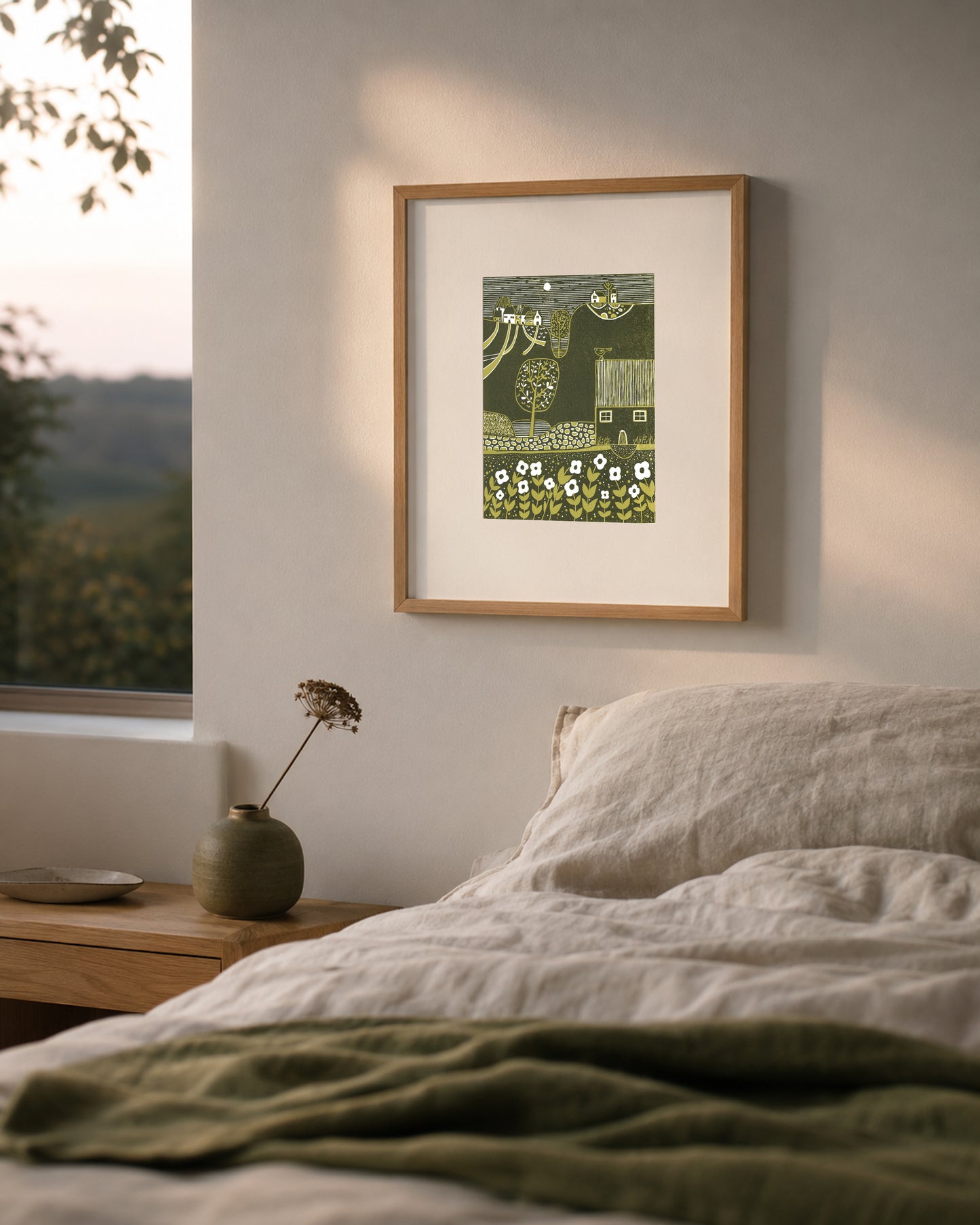 Framed Serene Hillside linocut print displayed above a bed in a softly lit bedroom with green textiles.