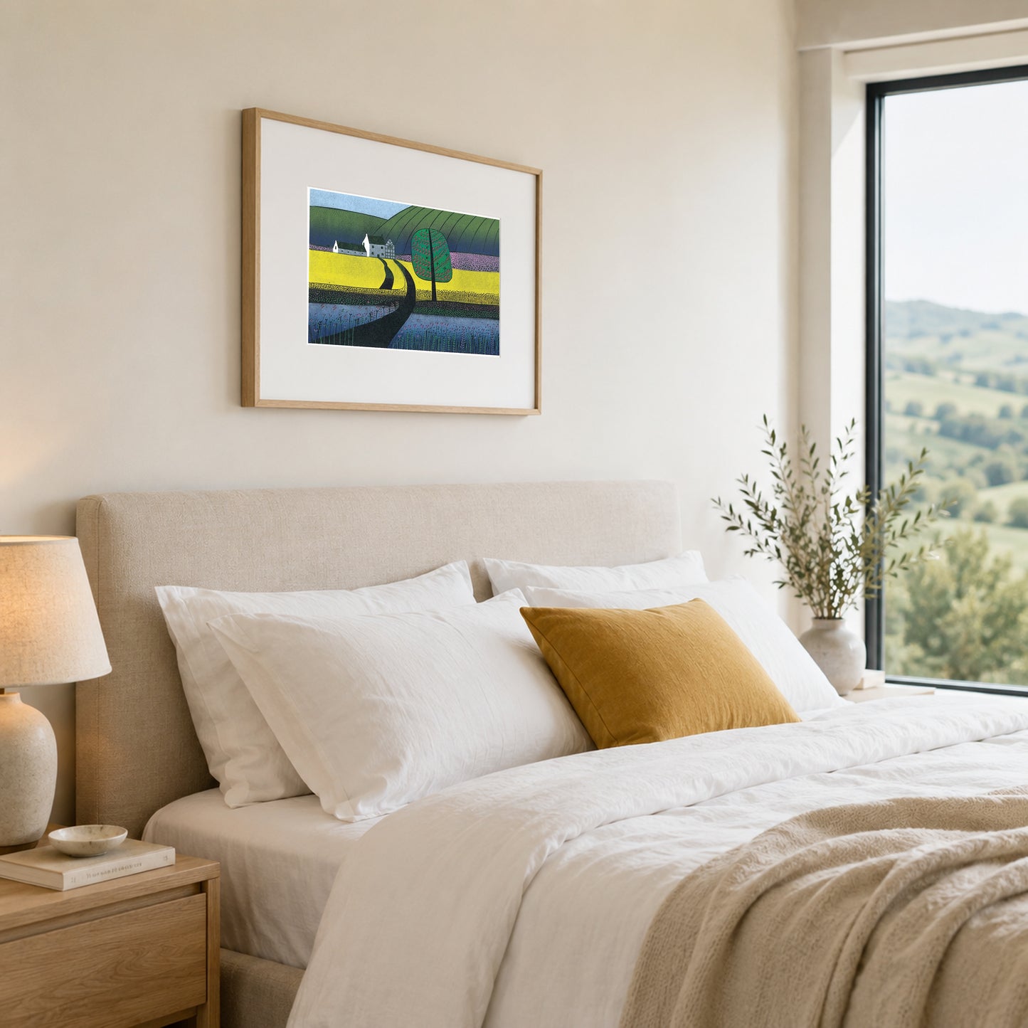 Framed landscape print on a warm cream wall above an upholstered headboard with a mustard cushion.