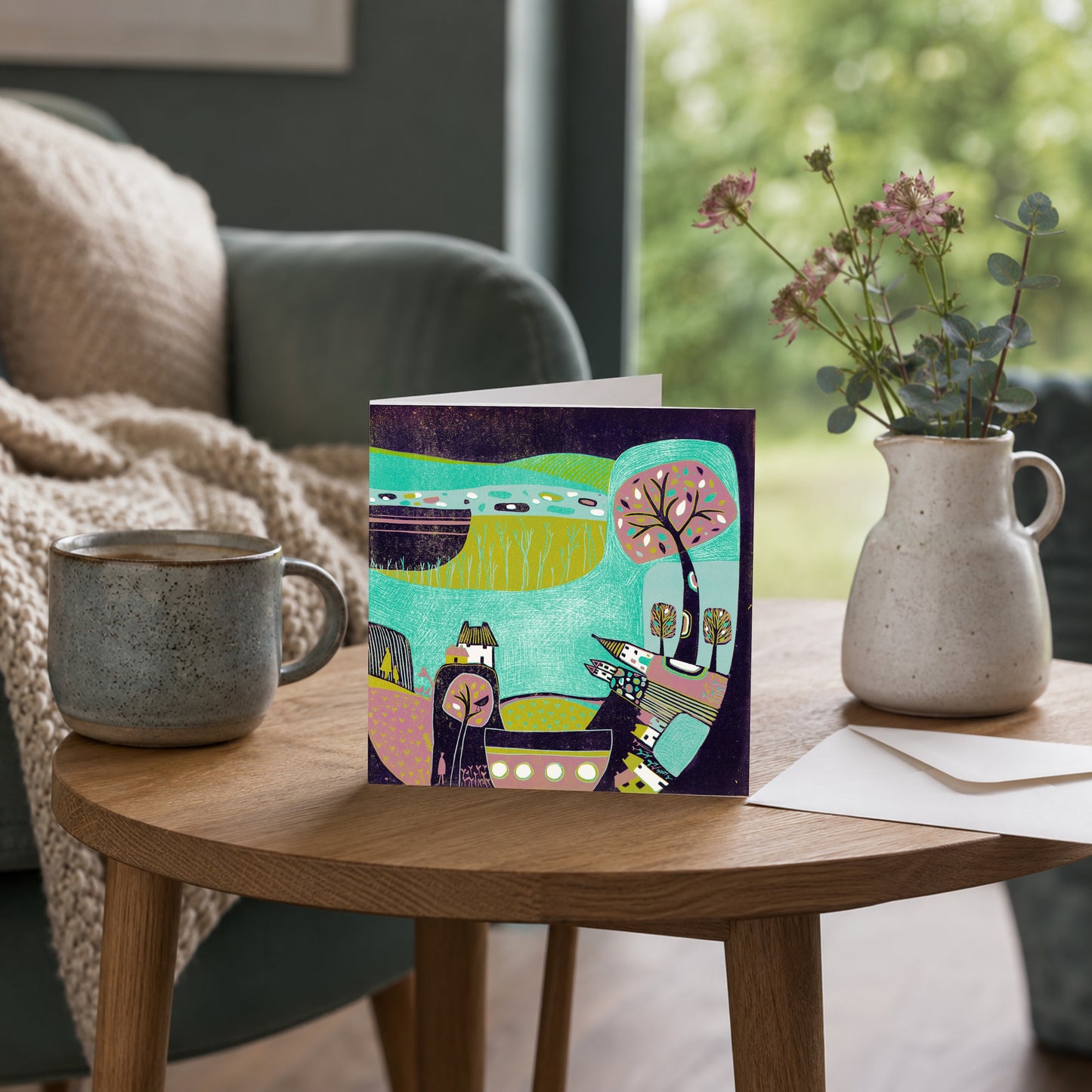 Linocut folk village greeting card propped against a mug on a table with a stone jug of flowers