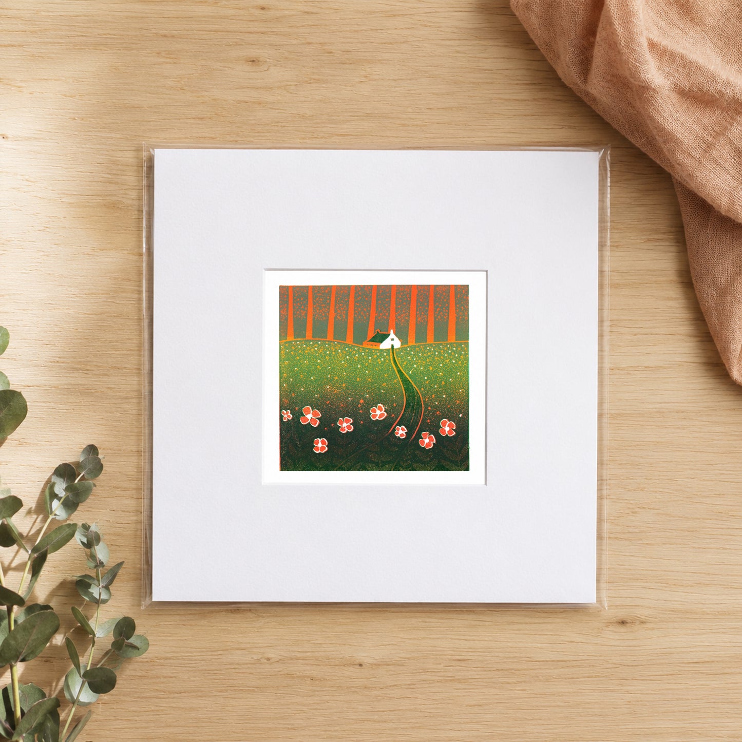 Mini linocut poppy meadow print in a white window mount, photographed flat on warm oak with eucalyptus