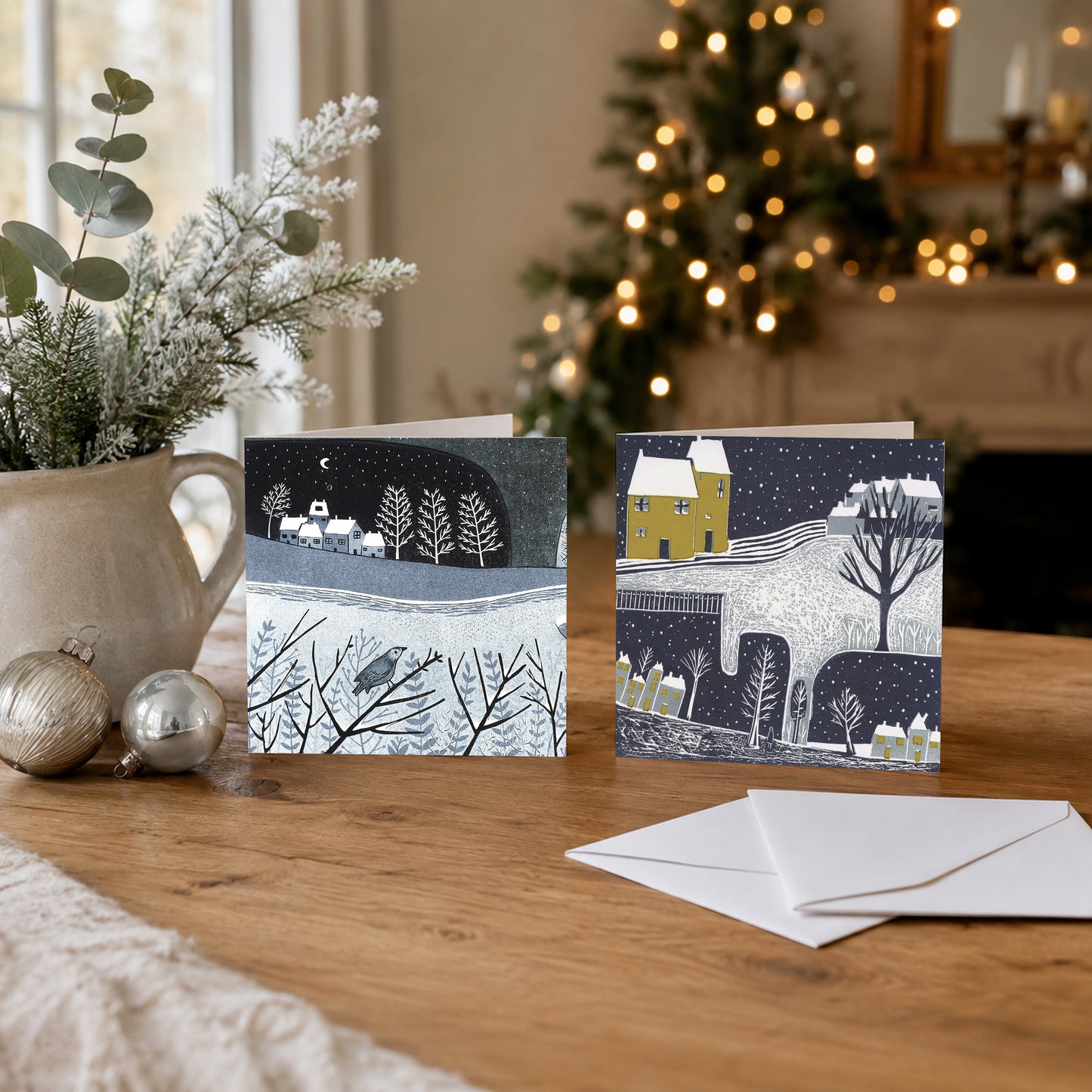 Two Wintry Nights Christmas cards with snowy linocut landscape designs displayed beside a Christmas tree and envelopes.