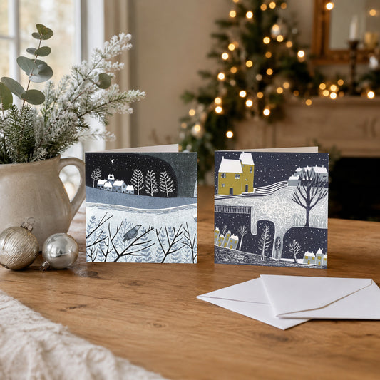 Two Wintry Nights Christmas cards with snowy linocut landscape designs displayed beside a Christmas tree and envelopes.