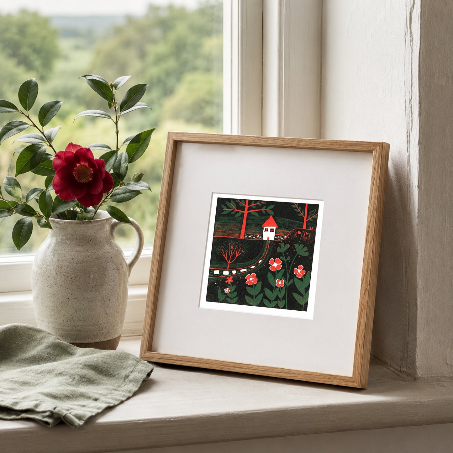 Mini linocut print framed in pale oak, propped on a windowsill with a red rose in a stone jug