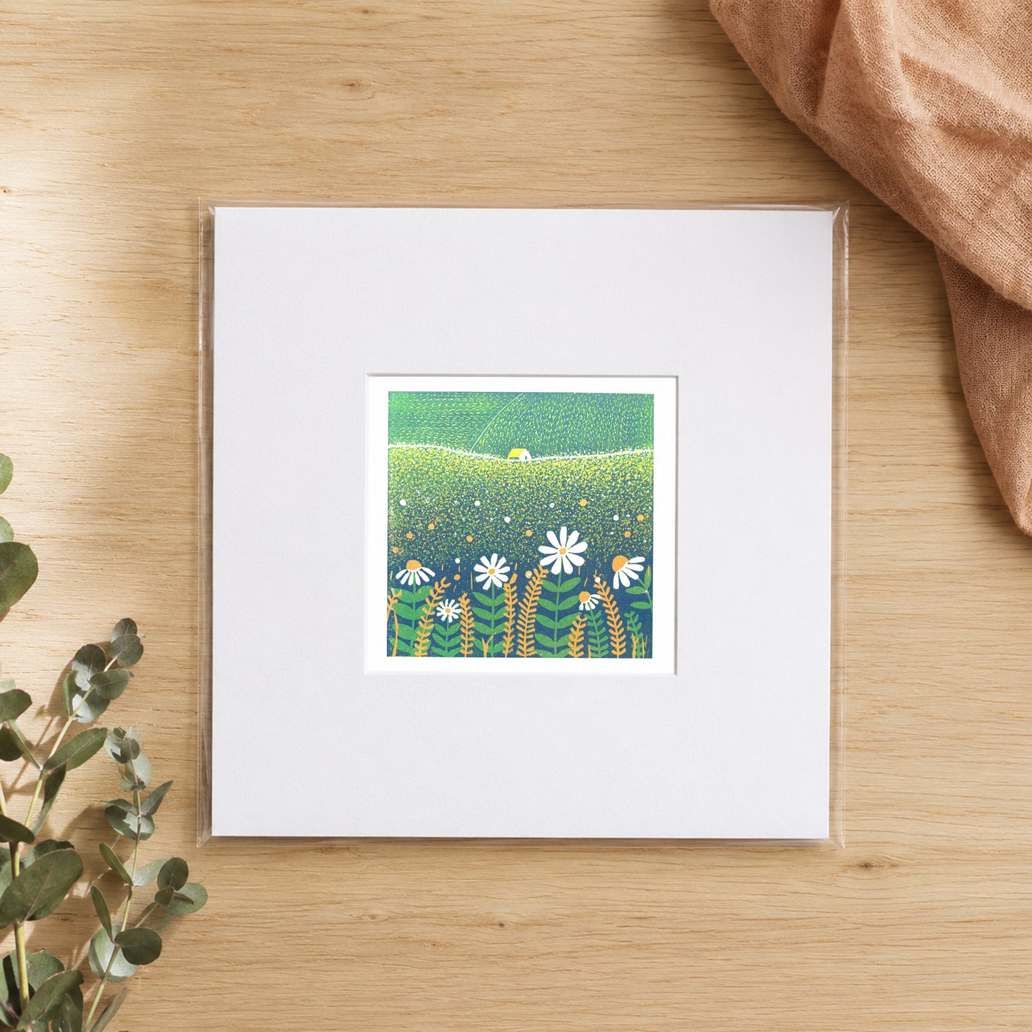 Mini linocut daisy field print in a white window mount, photographed flat on warm oak with eucalyptus