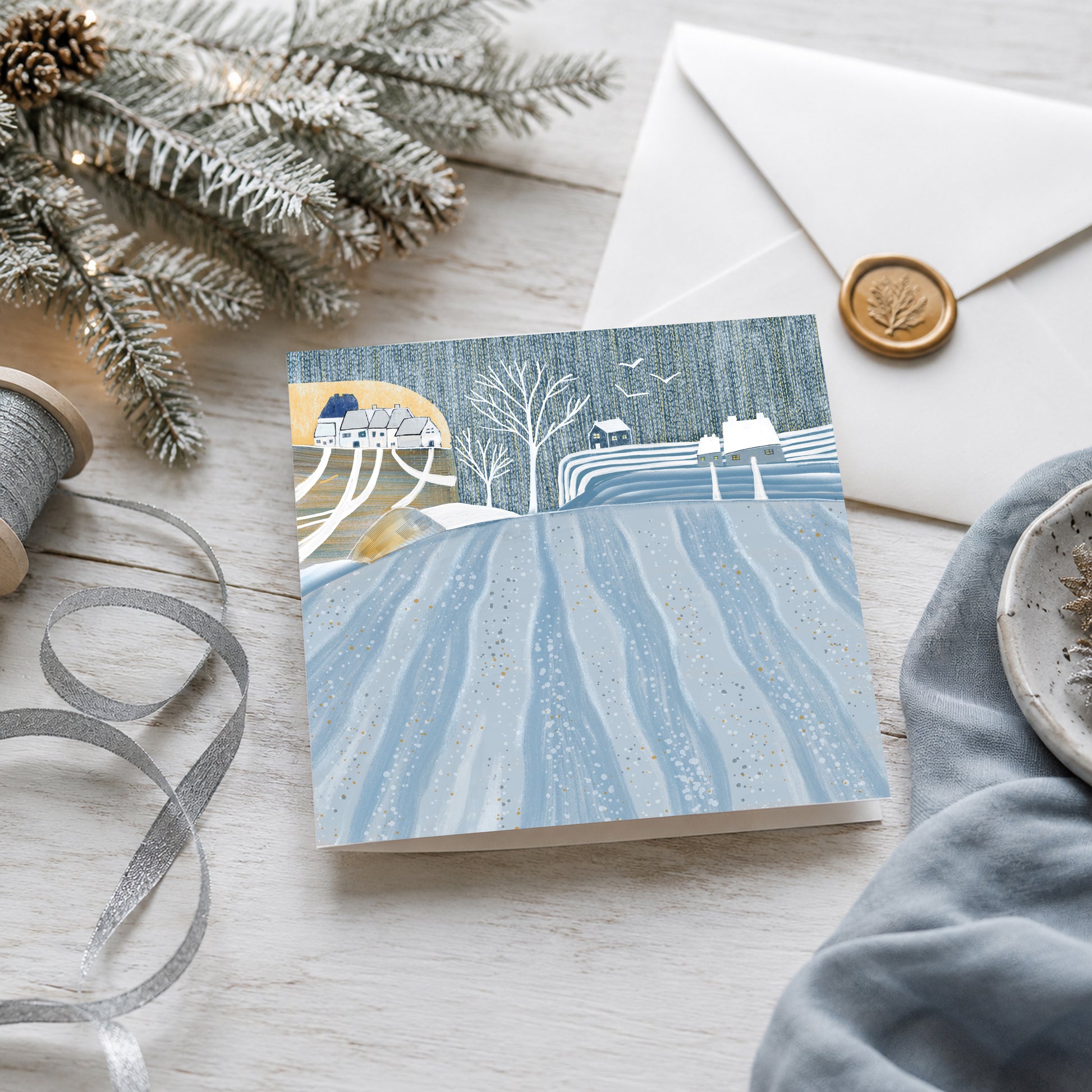 Winter Fields Christmas card featuring a snowy blue countryside landscape with cottages, displayed with an envelope, ribbon and frosted festive greenery.