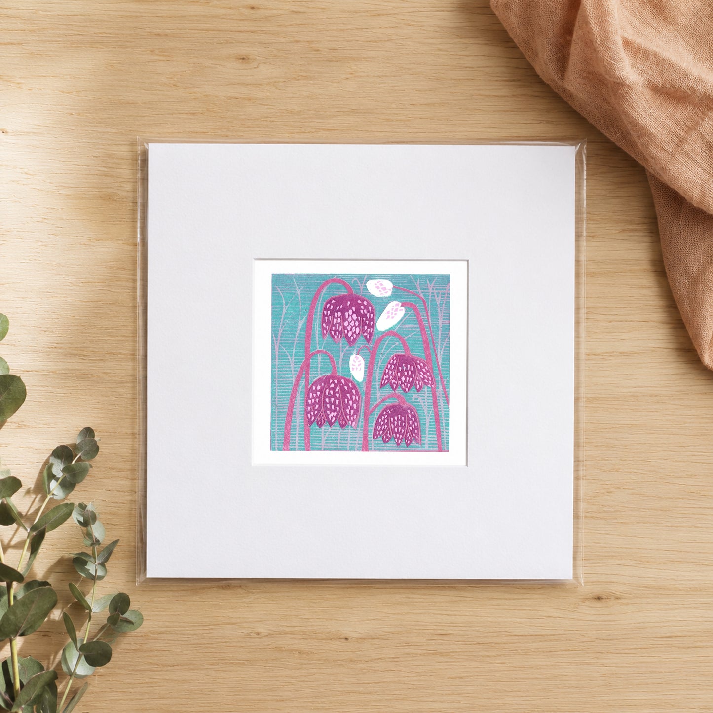 Mini linocut fritillary print in a white window mount, photographed flat on warm oak with eucalyptus