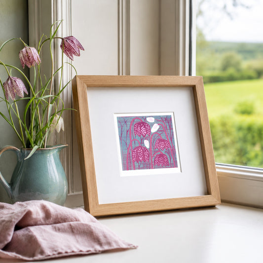 Mini linocut print framed in pale oak, propped on a windowsill with pink tulips in a stone vase