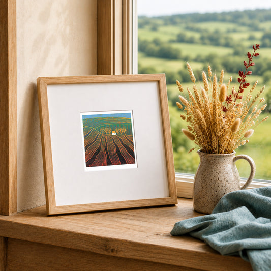 Mini linocut print framed in pale oak, propped on a windowsill with dried grasses in a stone jug