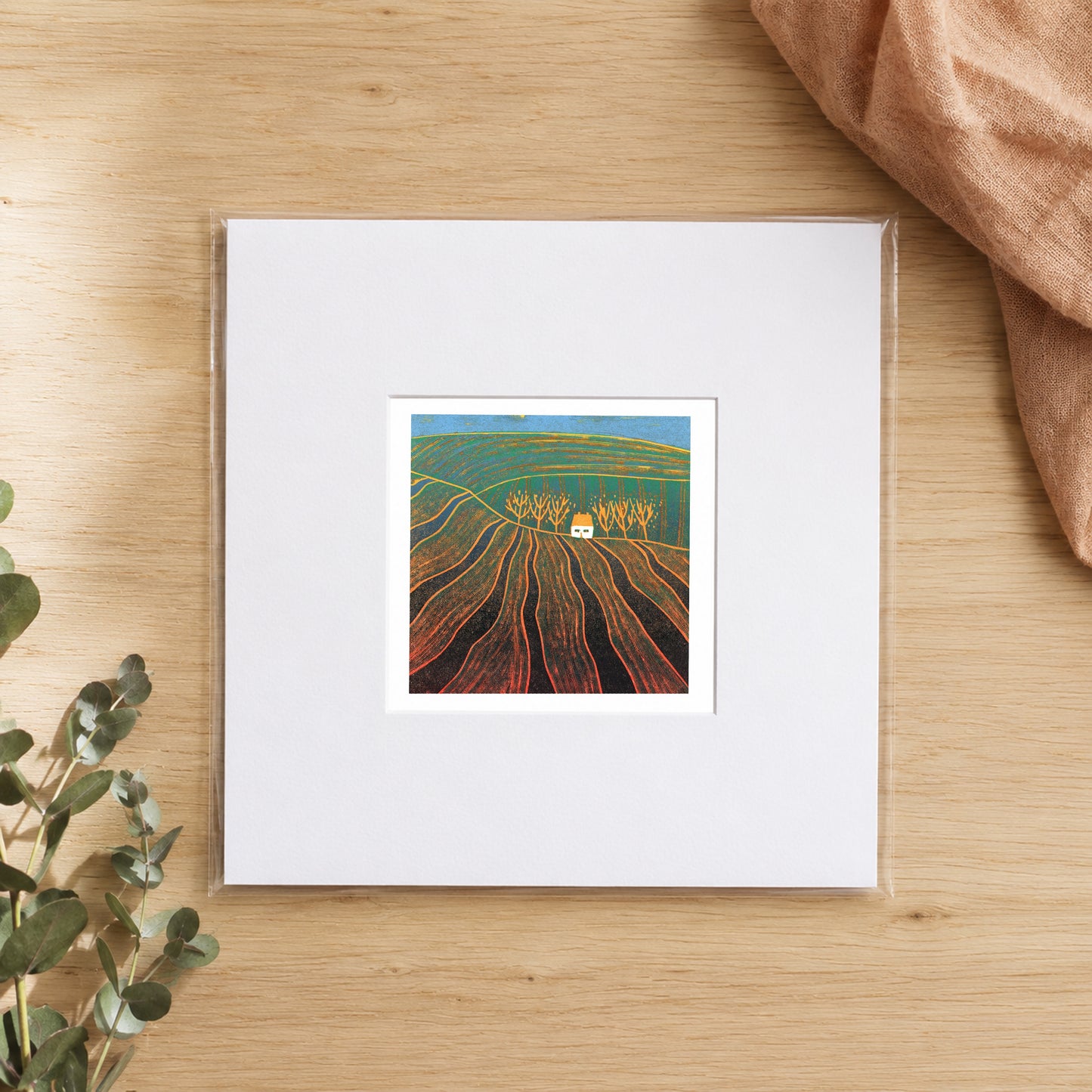 Mini linocut ploughed fields print in a white window mount, photographed flat on warm oak with eucalyptus