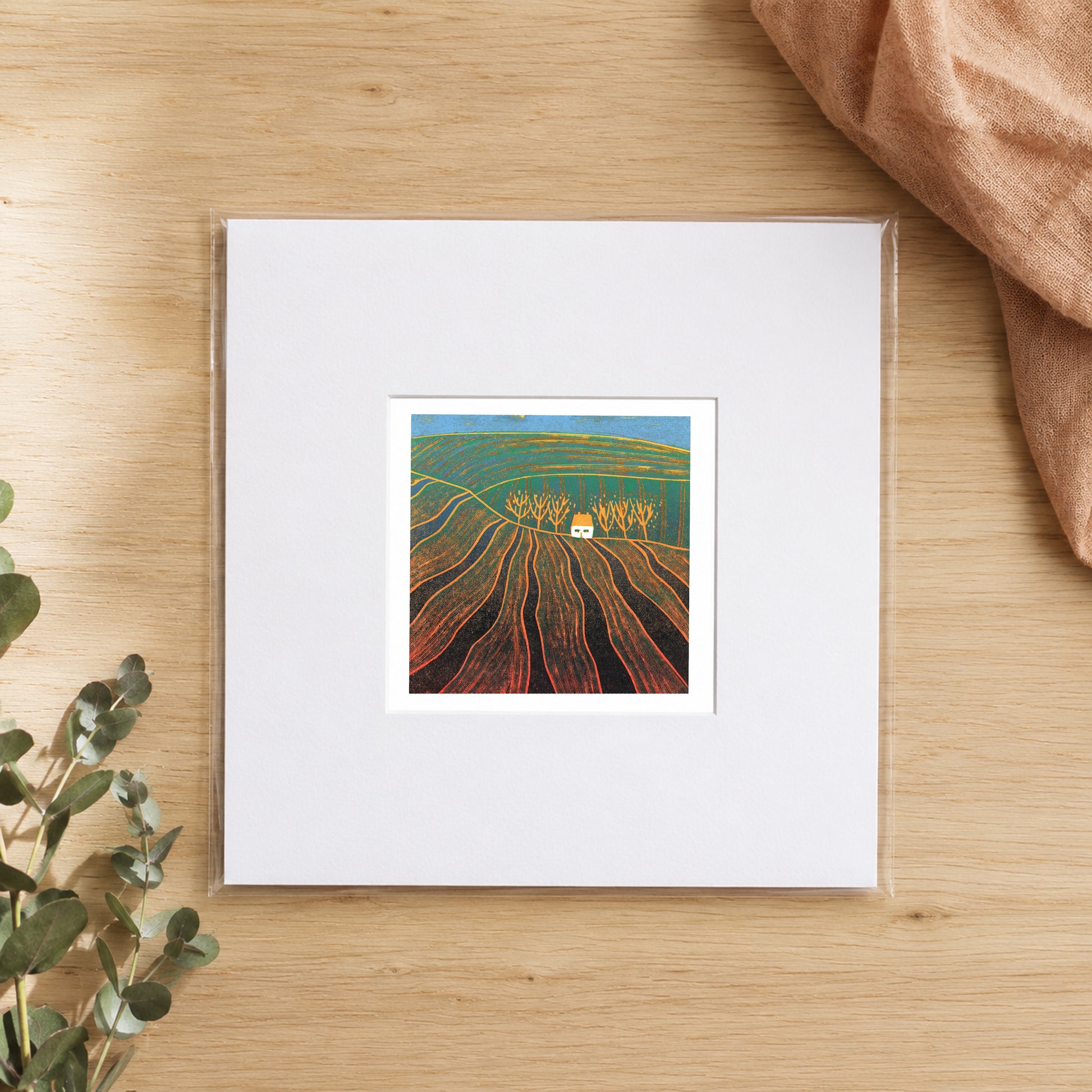 Mini linocut ploughed fields print in a white window mount, photographed flat on warm oak with eucalyptus