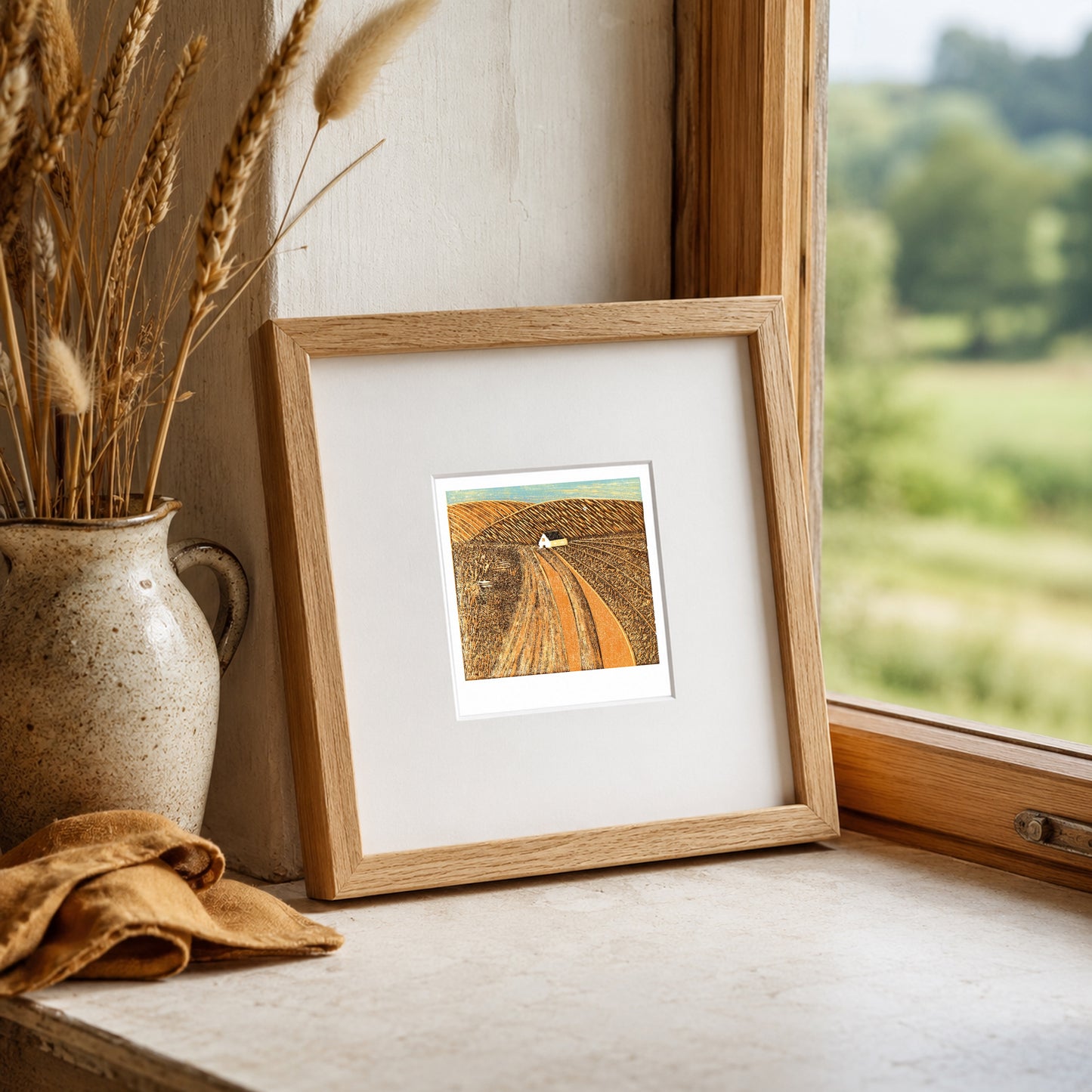 Mini linocut print framed in pale oak, propped on a windowsill with dried wheat in a stone vase