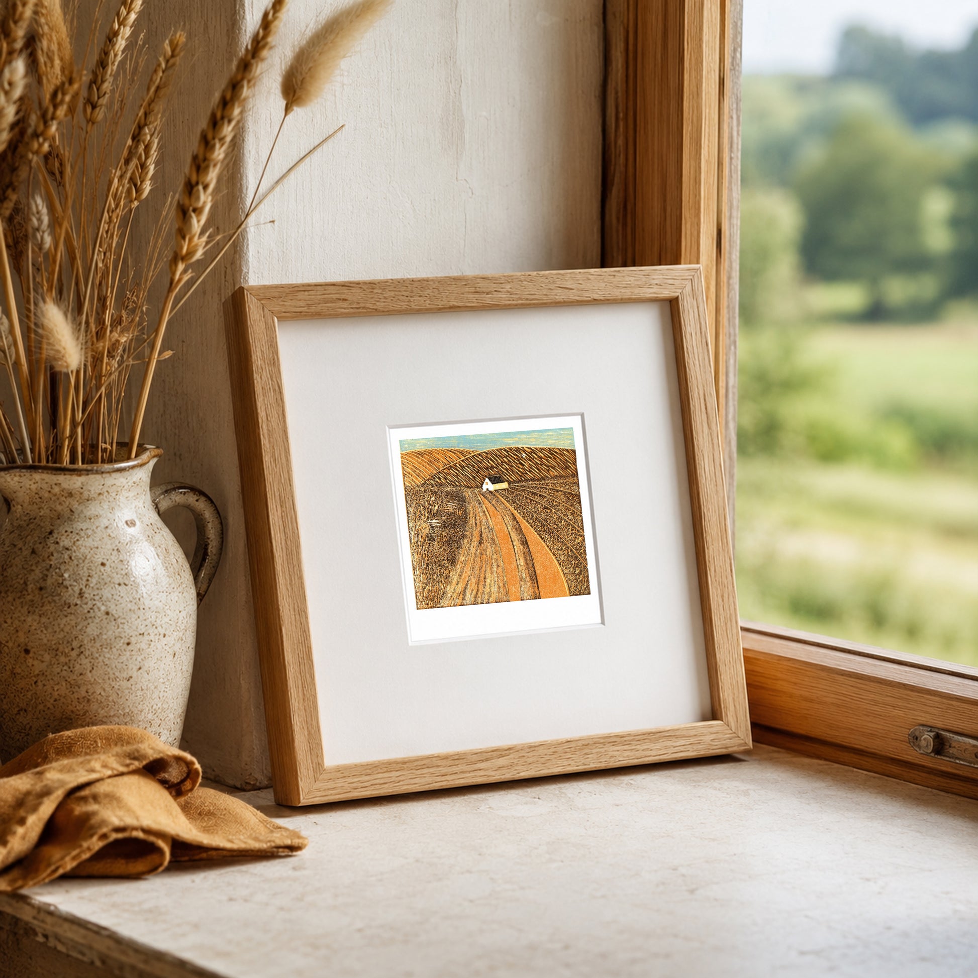 Mini linocut print framed in pale oak, propped on a windowsill with dried wheat in a stone vase