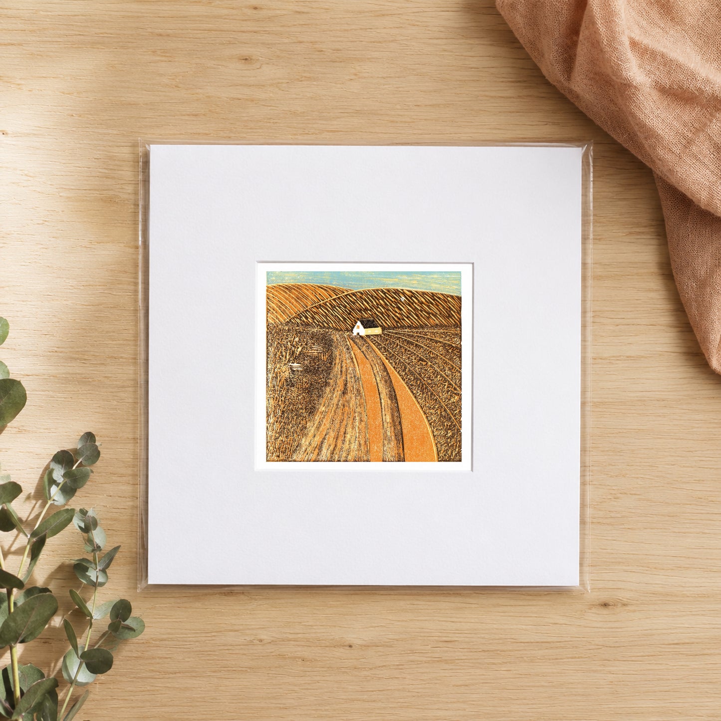 Mini linocut harvest fields print in a white window mount, photographed flat on warm oak with eucalyptus