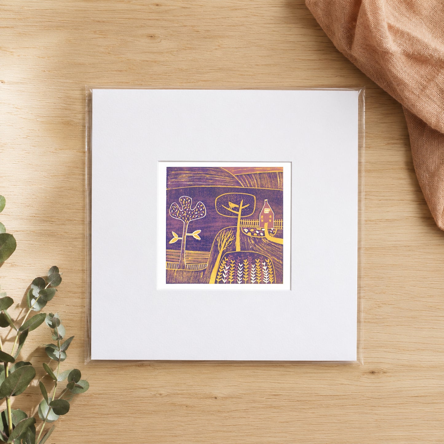 Mini linocut golden dusk print in a white window mount, photographed flat on warm oak with eucalyptus