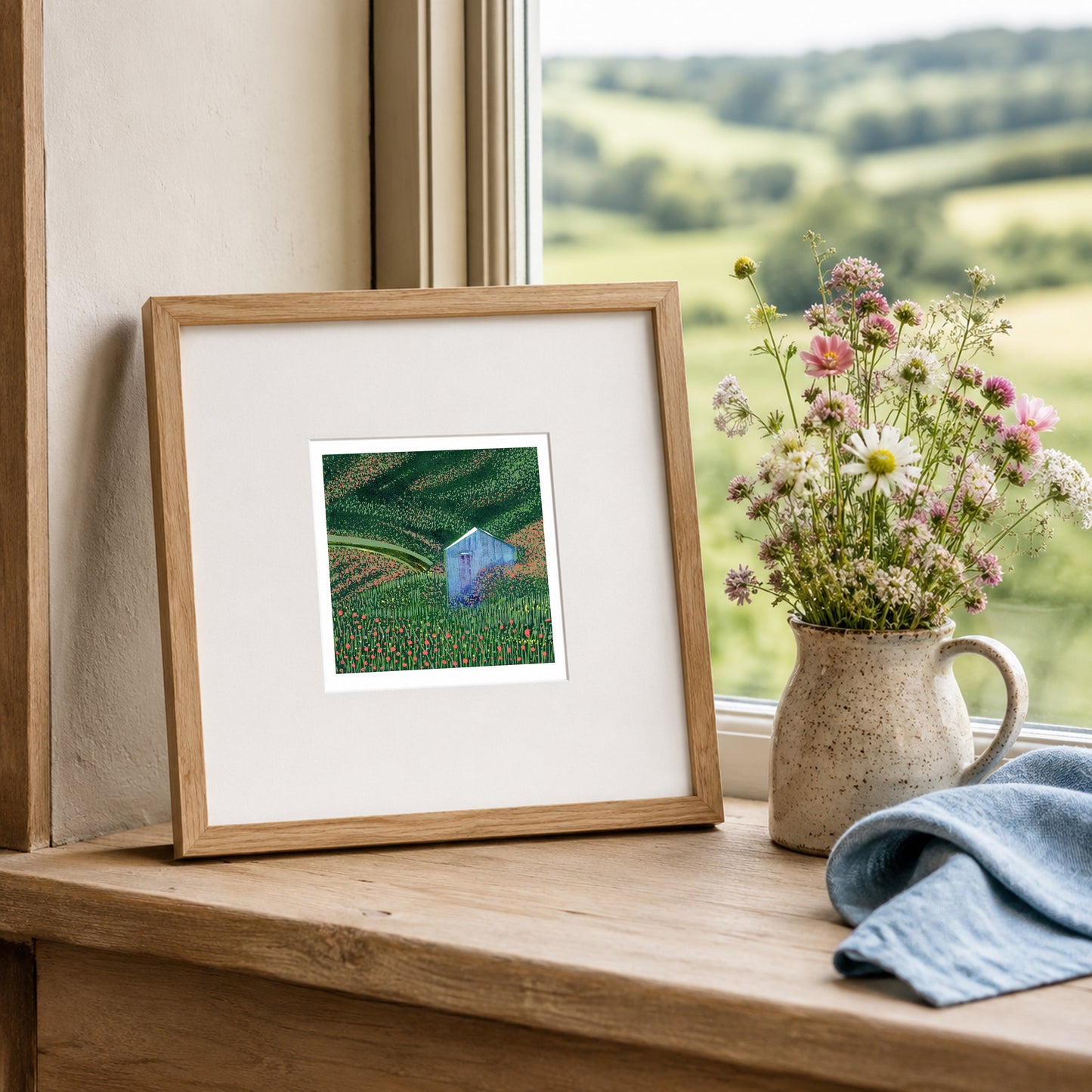 Mini linocut print framed in pale oak, propped on a windowsill with dried stems in a stone jug