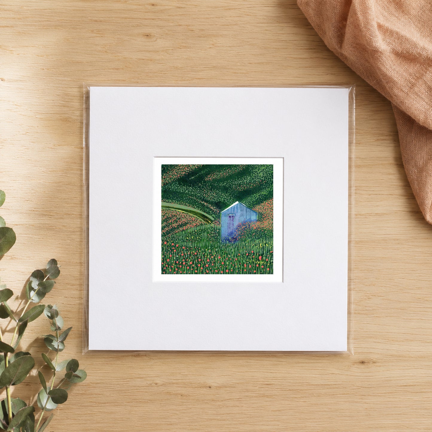 Mini linocut blue shed print in a white window mount, photographed flat on warm oak with eucalyptus