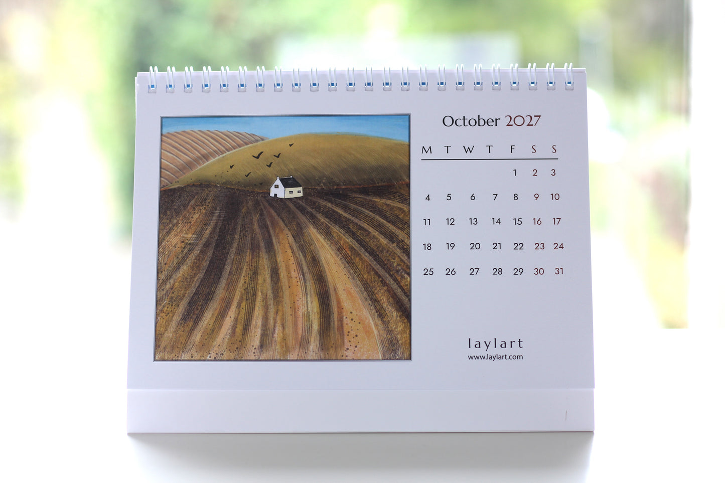 Photograph of the October 2027 desk calendar showing golden autumn fields, monthly dates, spiral binding and freestanding format.