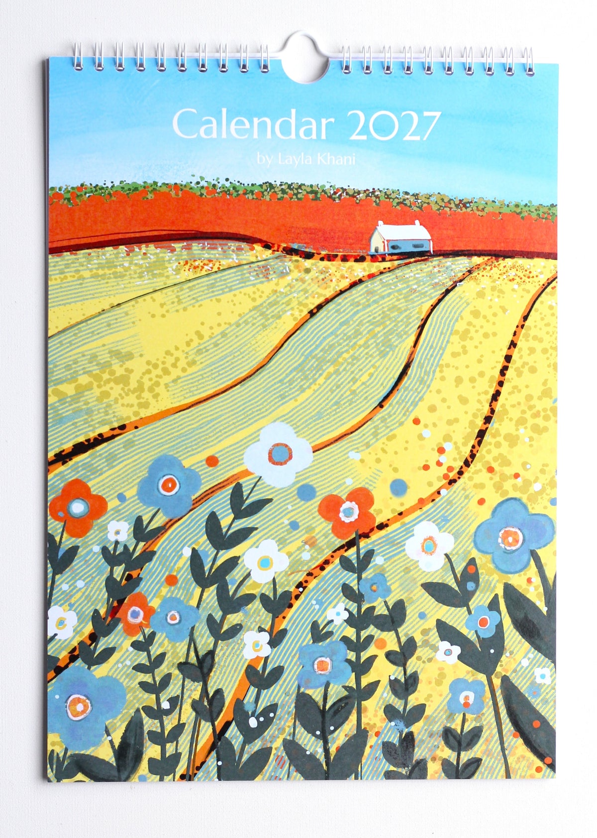 Photograph of the printed 2027 wall calendar cover showing yellow fields, flowers, spiral binding and hanging loop.