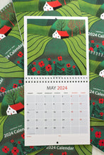 Load image into Gallery viewer, May 2024 calendar in 30 x 60 cm size, showcasing an open view of the entire calendar, featuring a picturesque linocut print of a rural landscape with a distant house, expansive lands, and vibrant red flowers in the foreground